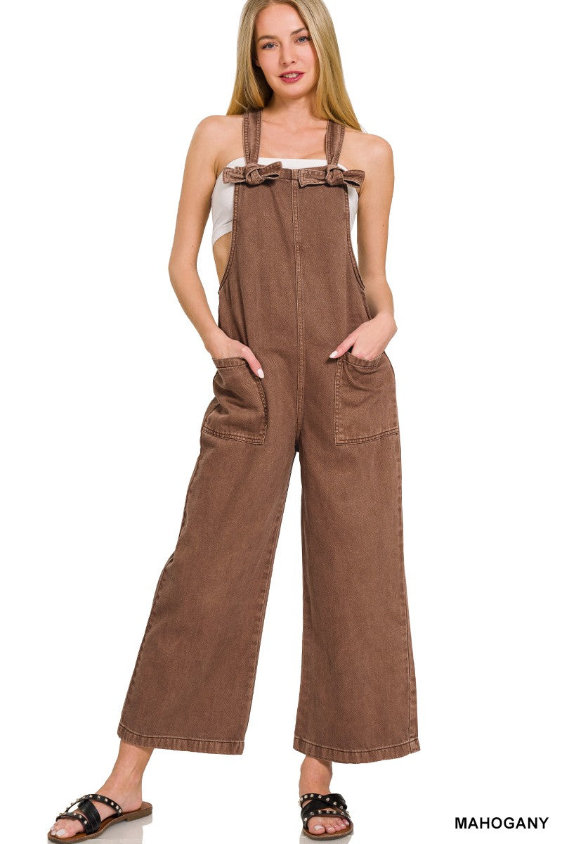 A female model wearing a brown washed knot strapped jumpsuit with pockets, paired with a white tank top and tan sandals.